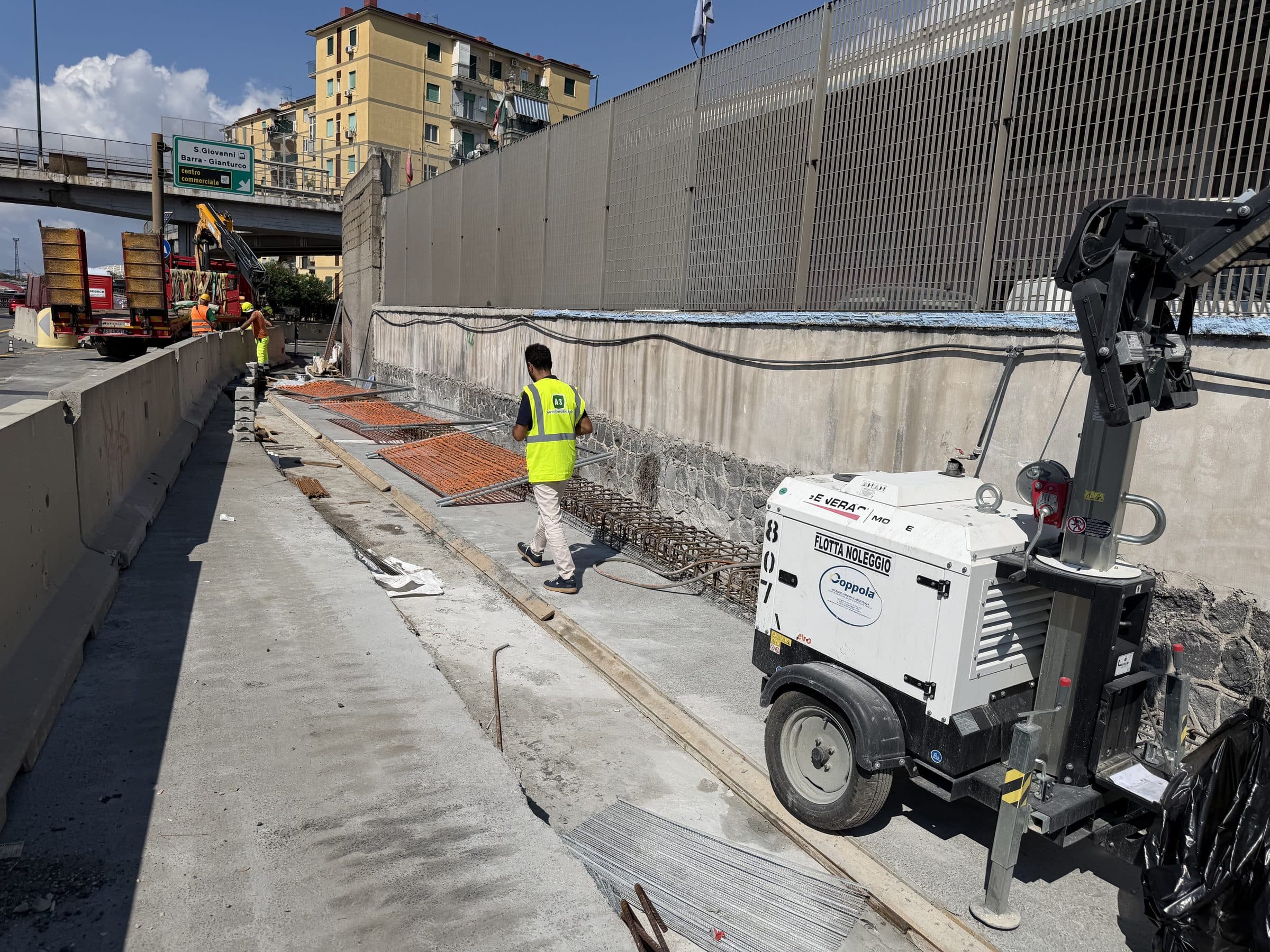 Corridoio di lavoro sotto alte barriere antirumore, con una torre faro e un operaio in abbigliamento ad alta visibilità