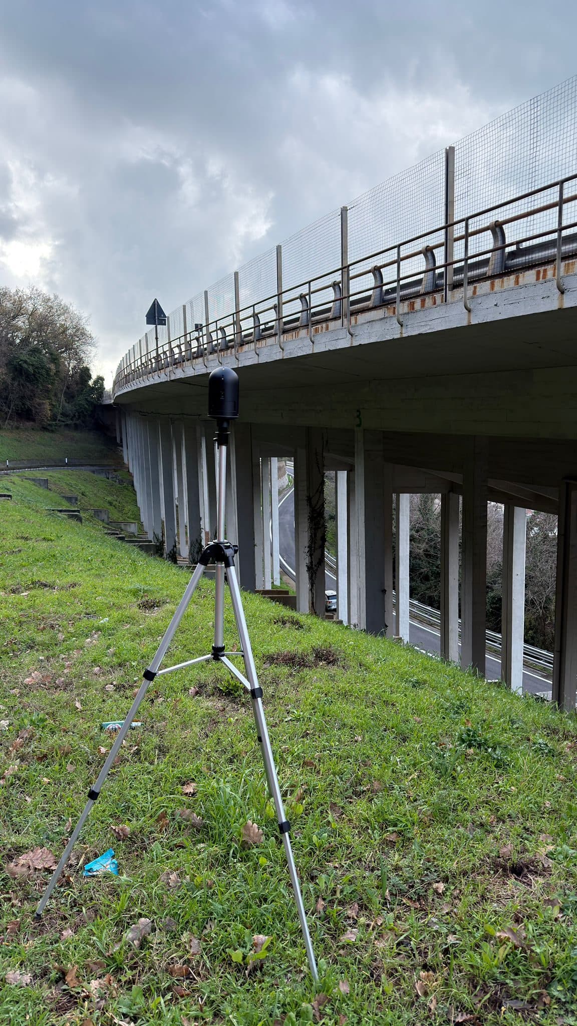 Laser scanner su treppiede nell’erba sotto l’impalcato di un viadotto sostenuto da pile snelle