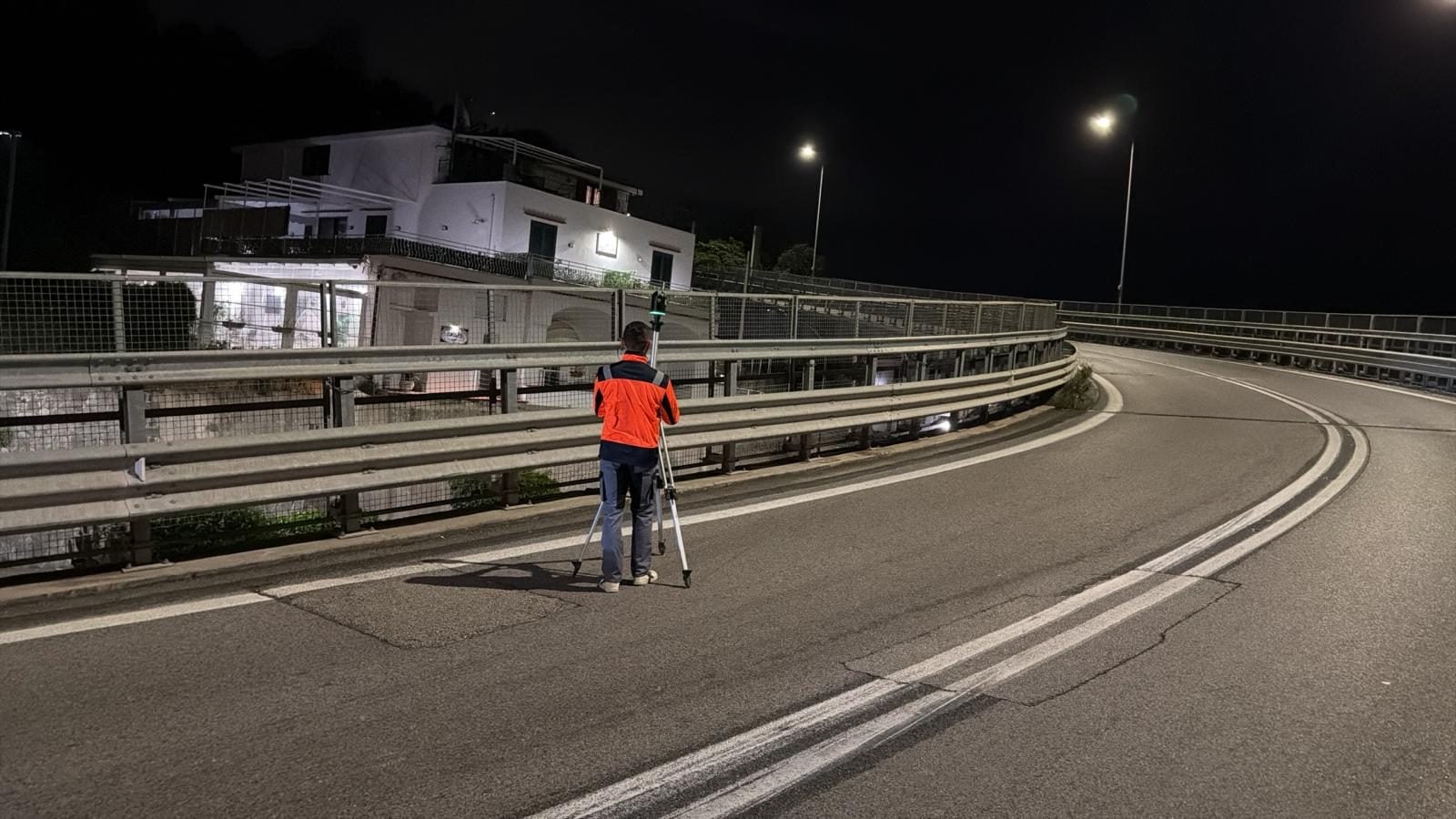 Tecnico in tuta arancione accanto a un laser scanner su treppiede, di notte, su una rampa in curva